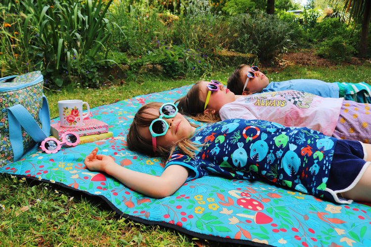 3 children lying on a mushromm print little lovebum picnic blanket in a park on the green grass