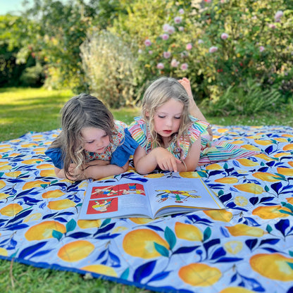 Two young girls lying on a grassy lawn reading a book on a colorful picnic blanket with a yellow and blue lemon pattern, with garden foliage in the background.