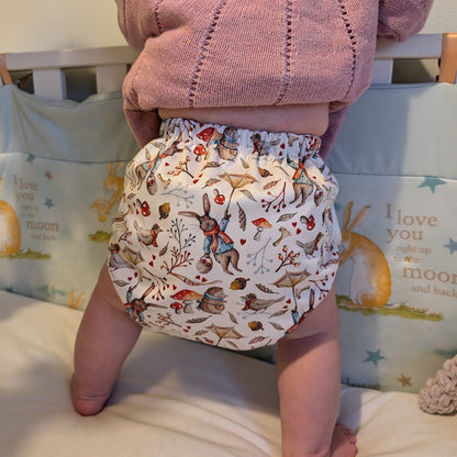 Baby wearing a pink knitted cardigan and patterned diaper standing on a bed.