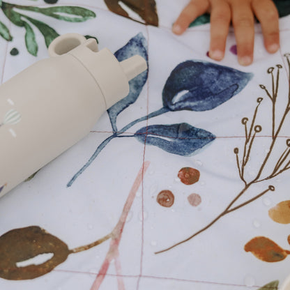 A close-up of a child's hand resting on a white quilted blanket with a watercolor botanical pattern, with a cream-colored bottle partially visible in the foreground.